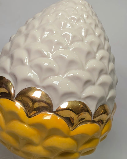 Pigna portafortuna in ceramica bicolore gialla e bianca lucida, impreziosita da una raffinata corona di squame dorate che ne definisce il profilo centrale. Questo elegante oggetto ornamentale artigianale poggia su una base circolare con finiture in oro, rappresentando un complemento d'arredo perfetto per decorazioni d'interni in stile chic e contemporaneo.