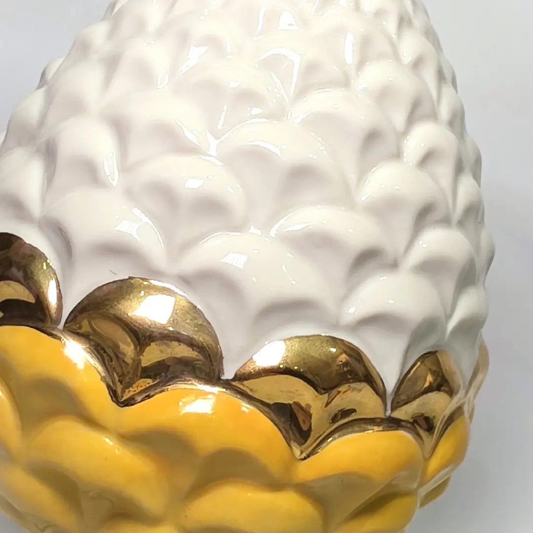 Pigna portafortuna in ceramica bicolore gialla e bianca lucida, impreziosita da una raffinata corona di squame dorate che ne definisce il profilo centrale. Questo elegante oggetto ornamentale artigianale poggia su una base circolare con finiture in oro, rappresentando un complemento d'arredo perfetto per decorazioni d'interni in stile chic e contemporaneo.