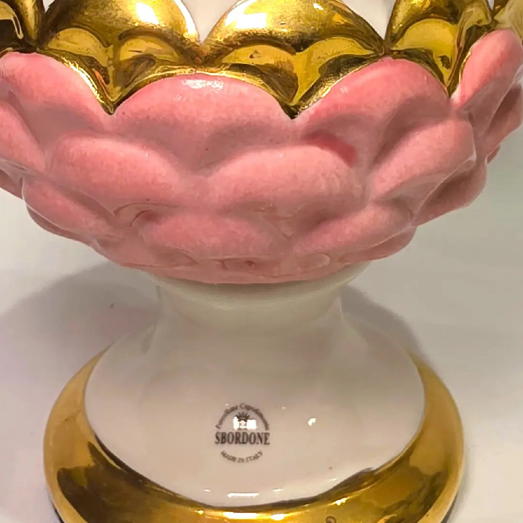 Pigna portafortuna in ceramica bicolore rosa e bianca lucida, impreziosita da una raffinata corona di squame dorate che ne definisce il profilo centrale. Questo elegante oggetto ornamentale artigianale poggia su una base circolare con finiture in oro, rappresentando un complemento d'arredo perfetto per decorazioni d'interni in stile chic e contemporaneo.