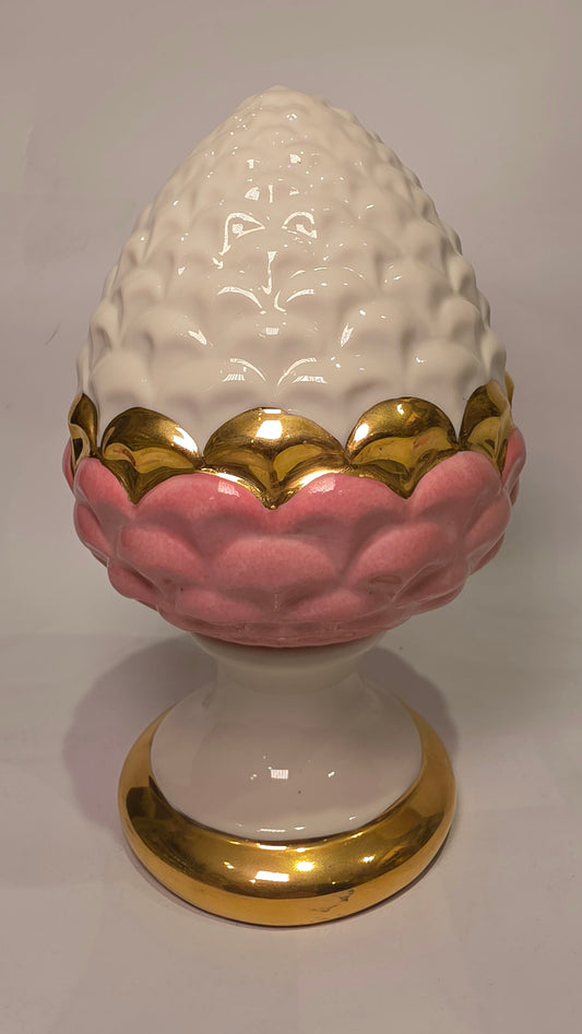 Pigna portafortuna in ceramica bicolore rosa e bianca lucida, impreziosita da una raffinata corona di squame dorate che ne definisce il profilo centrale. Questo elegante oggetto ornamentale artigianale poggia su una base circolare con finiture in oro, rappresentando un complemento d'arredo perfetto per decorazioni d'interni in stile chic e contemporaneo.