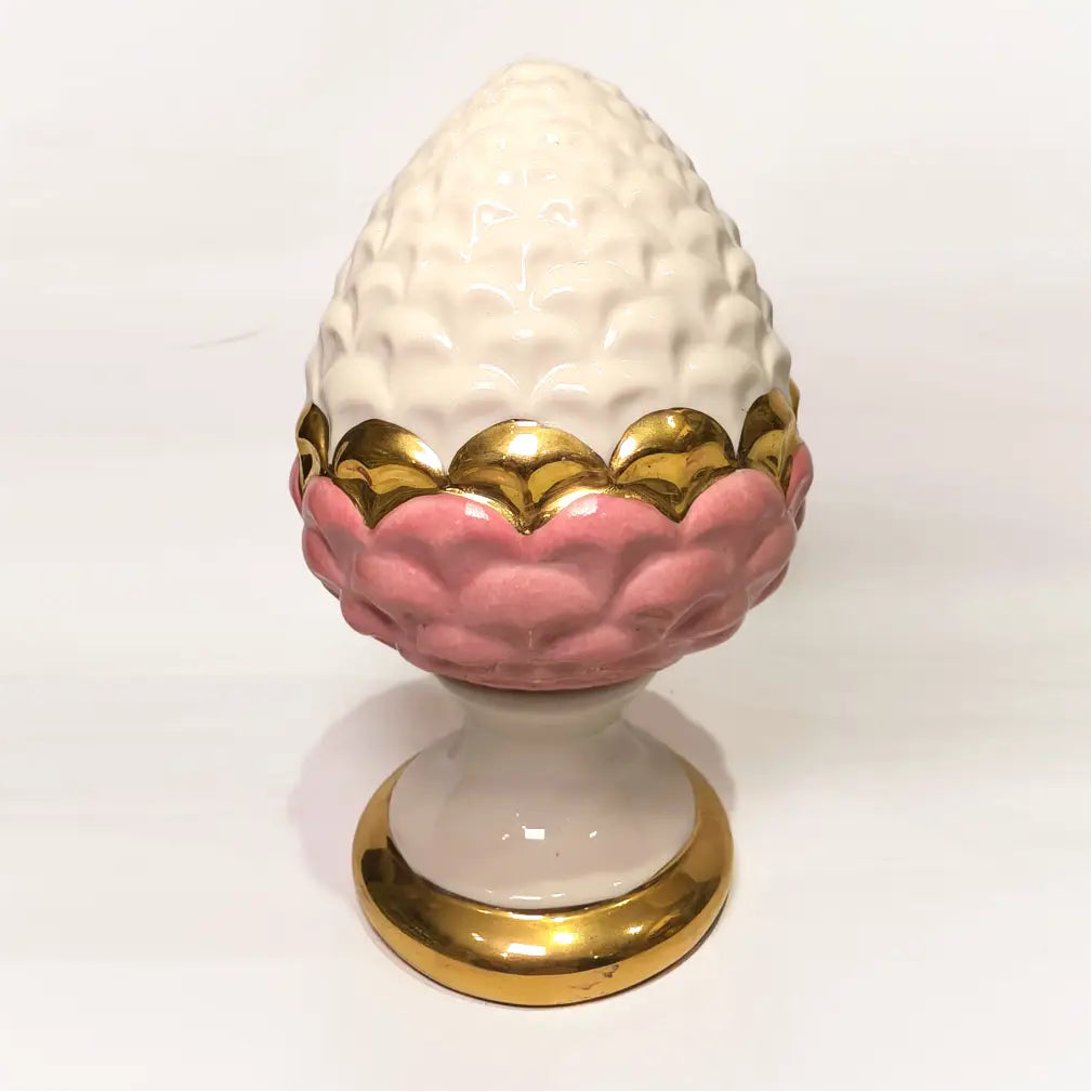 Pigna portafortuna in ceramica bicolore rosa e bianca lucida, impreziosita da una raffinata corona di squame dorate che ne definisce il profilo centrale. Questo elegante oggetto ornamentale artigianale poggia su una base circolare con finiture in oro, rappresentando un complemento d'arredo perfetto per decorazioni d'interni in stile chic e contemporaneo.