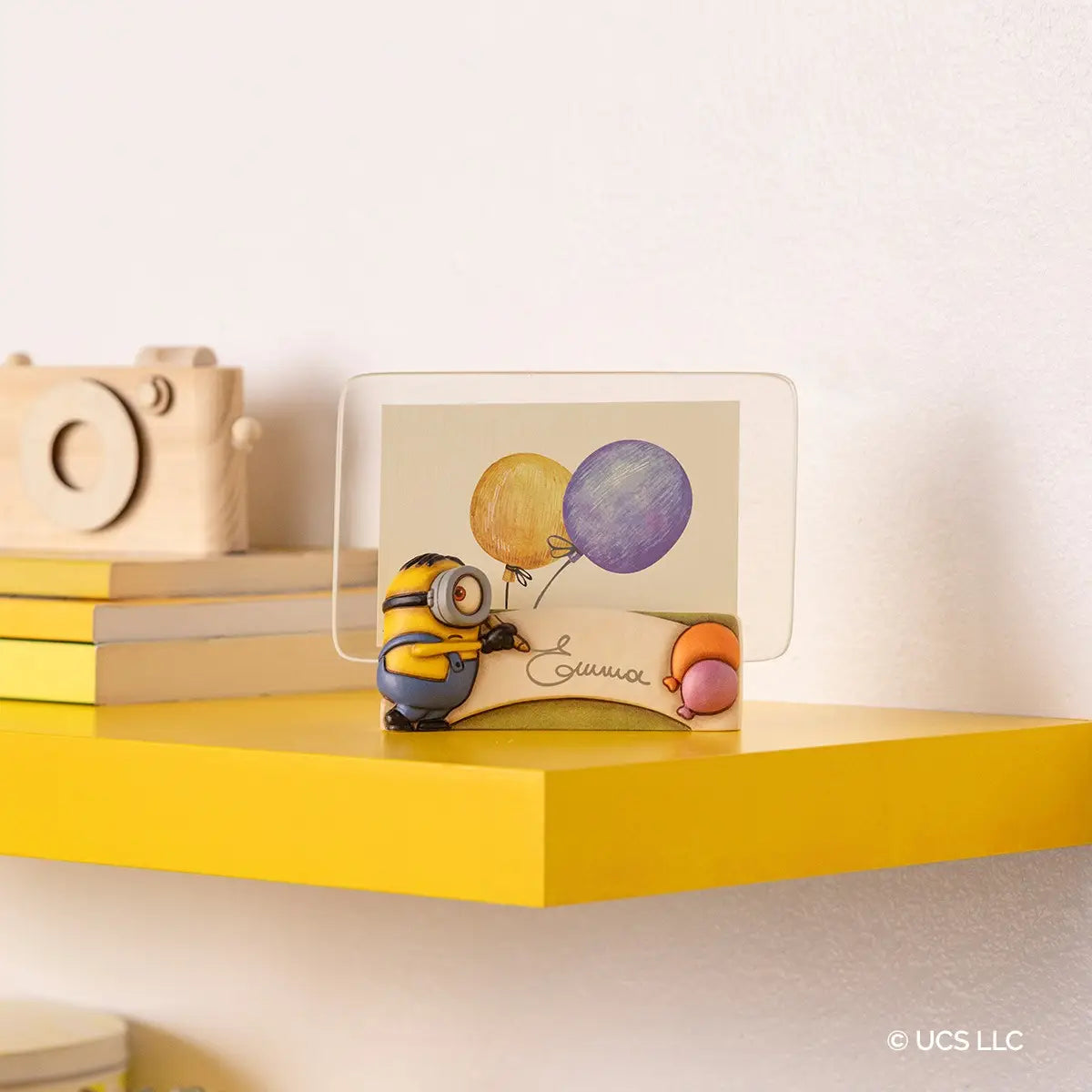 Cornice portafoto in ceramica Thun con personaggio Minion in rilievo. La base della cornice presenta un Minion con salopette azzurra intento a scrivere su una pergamena bianca, affiancato da due palloncini colorati. Design iconico Thun applicato al mondo dei Minions, ideale per camerette o come idea regalo originale.