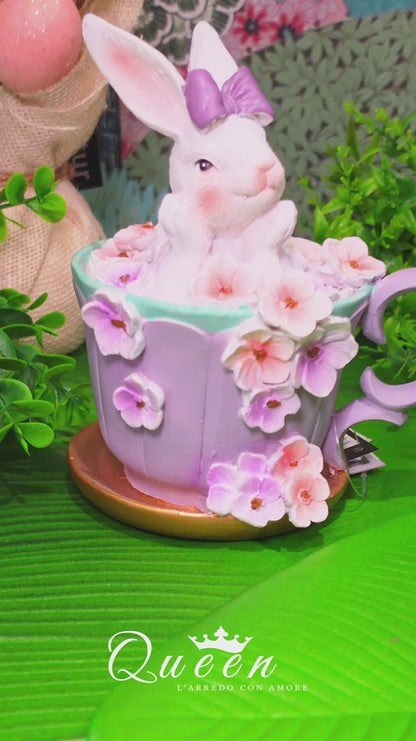Decorazione pasquale scultorea Vetur in poliresina. Coniglietto bianco con fiocco viola seduto all'interno di una tazza da tè lilla decorata con fiori rosa e viola sfumati, appoggiata su un piattino dorato.