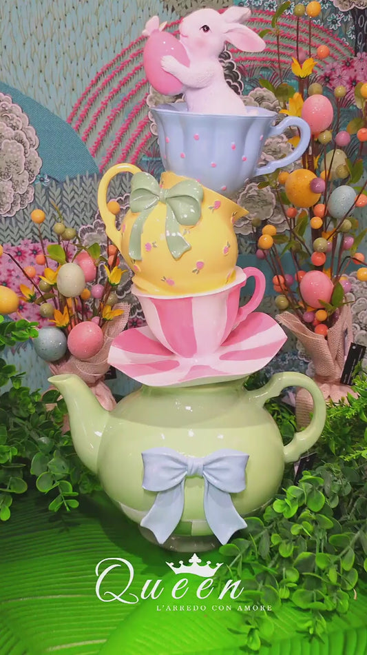 Statuina decorativa in resina composta da una torre di stoviglie multicolor: alla base una teiera verde con fiocco azzurro, sormontata da un piattino, una tazzina a righe rosa, una brocca gialla con fiocco verde e una tazzina azzurra a pois. In cima, un coniglietto bianco sbuca dalla tazzina tenendo tra le zampe un uovo di Pasqua rosa. Design verticale dai colori pastello.
