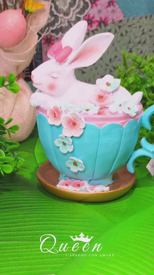 Decorazione pasquale scultorea Vetur in poliresina. Coniglietto bianco con fiocco rosaseduto all'interno di una tazza da tè azzurra decorata con fiori rosa e celesti sfumati, appoggiata su un piattino dorato.