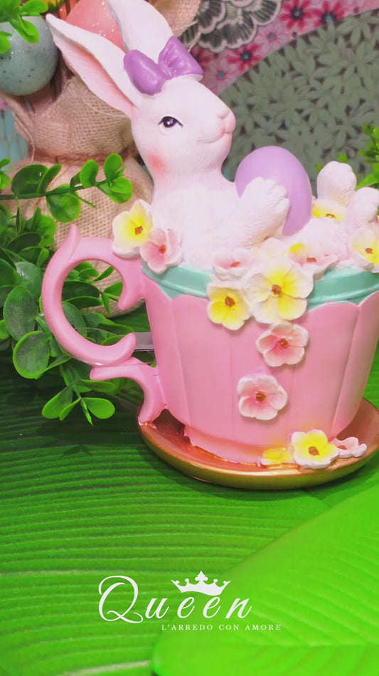 Decorazione pasquale Vetur in poliresina. Coniglietto bianco con fiocco viola seduto in una tazza rosa decorata con fiori gialli e bianchi, mentre regge un uovo viola. Altezza 16,5 cm.