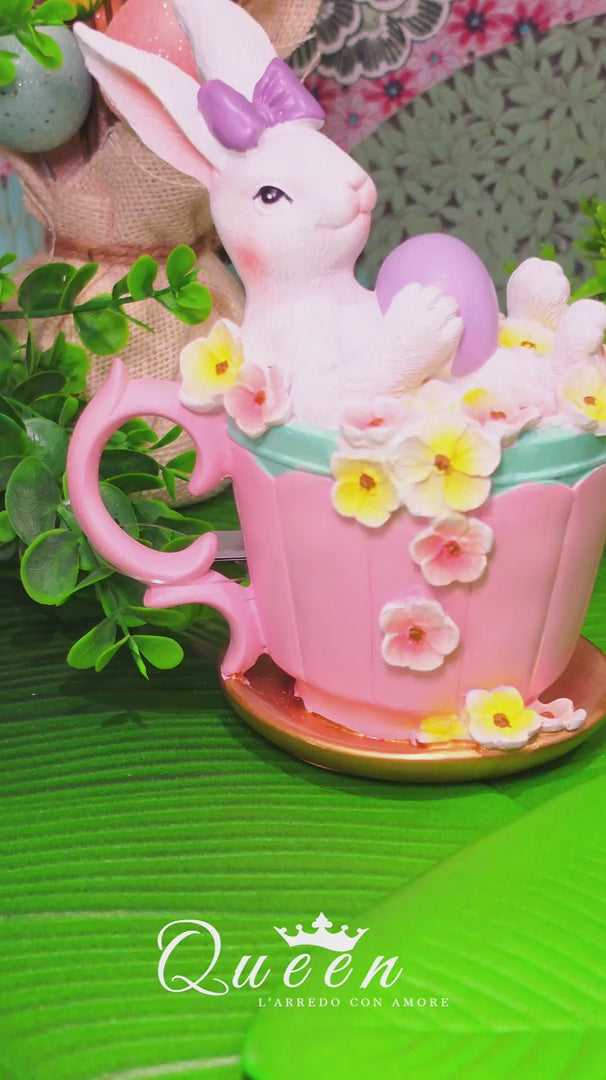 Decorazione pasquale Vetur in poliresina. Coniglietto bianco con fiocco viola seduto in una tazza rosa decorata con fiori gialli e bianchi, mentre regge un uovo viola. Altezza 16,5 cm.