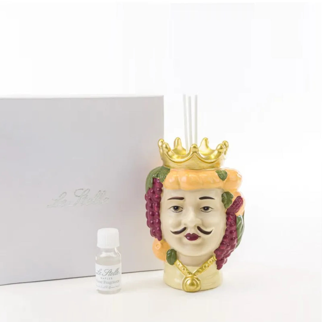 Profumatore d'ambiente in ceramica dipinta a mano raffigurante una testa di moro maschile con corona dorata, decorata con grappoli d'uva e agrumi colorati, con bastoncini bianchi e boccetta di fragranza "Home Fragrance". Diffusore dalla tradizione siciliana firmato Le Stelle, presentato con la sua confezione bianca del marchio, ideale come idea regalo raffinata.