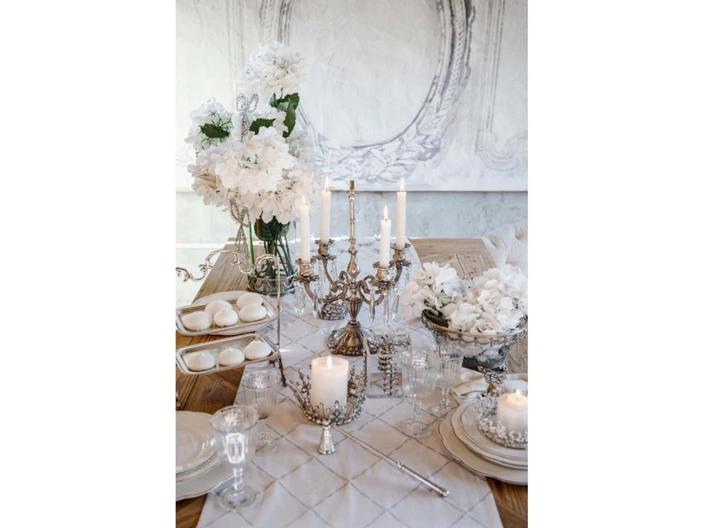Tavola apparecchiata con runner bianco Blanc Mariclò a motivi di fiocchi argento. La mise en place include candelabri classici, alzatine con meringhe e fiori bianchi per uno stile shabby sofisticato.