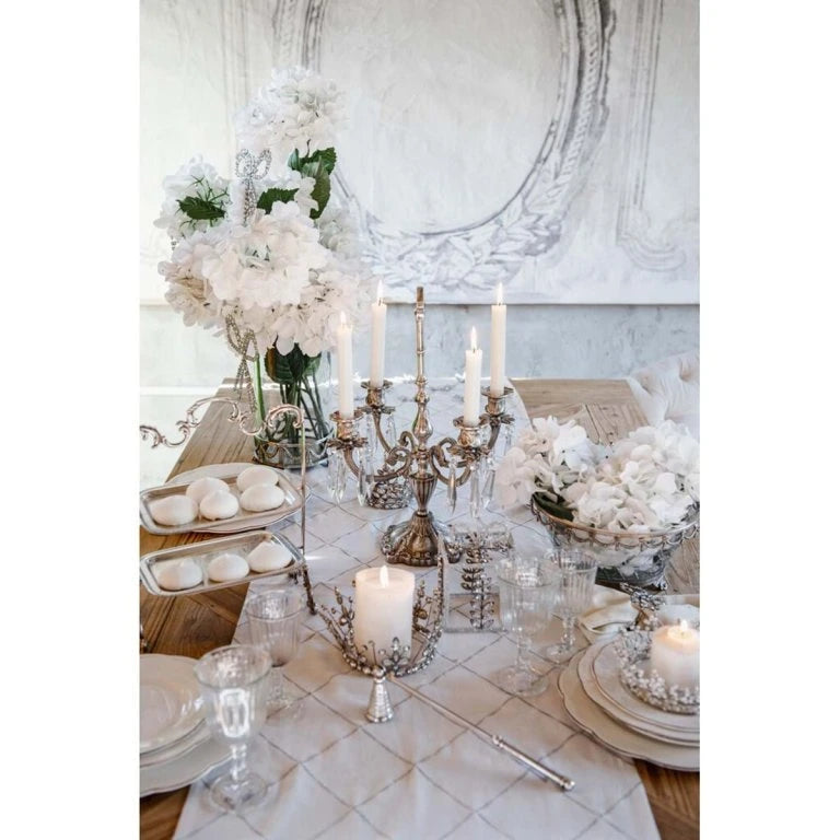 Tavola apparecchiata con runner bianco Blanc Mariclò a motivi di fiocchi argento. La mise en place include candelabri classici, alzatine con meringhe e fiori bianchi per uno stile shabby sofisticato.