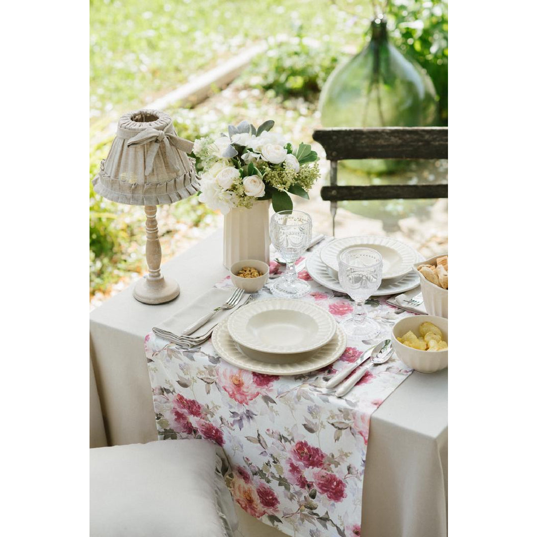 Tavola apparecchiata all'aperto con runner floreale rosa e beige. Mise en place elegante con piatti in ceramica crema, calici in vetro lavorato, fiori bianchi e una piccola lampada in tessuto Shabby.