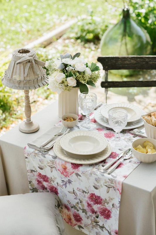 Tavola apparecchiata all'aperto con runner floreale rosa e beige. Mise en place elegante con piatti in ceramica crema, calici in vetro lavorato, fiori bianchi e una piccola lampada in tessuto Shabby.