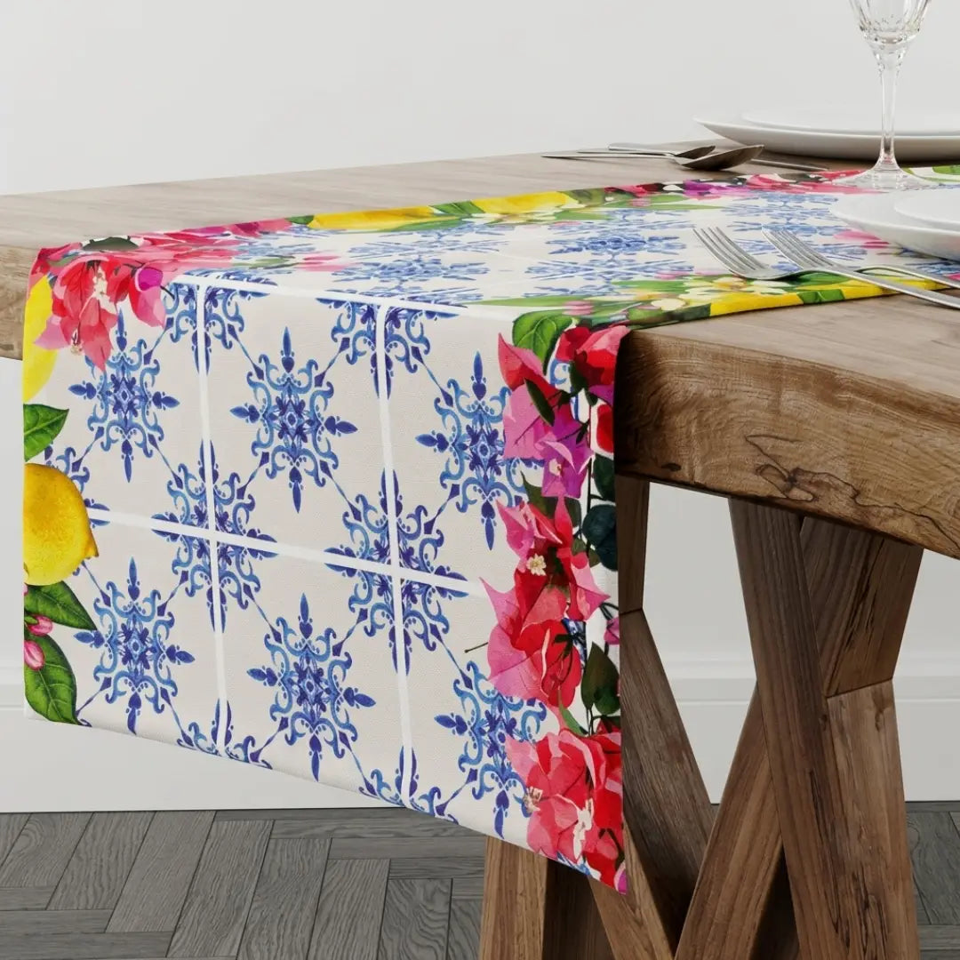 Runner con maioliche blu e fiori caratterizzato da un elegante motivo a piastrelle geometriche in stile mediterraneo su fondo bianco. I bordi di questo runner da tavola sono impreziositi da vivaci fiori rosa fucsia, limoni gialli e foglie verdi, ideali per decorare la tavola con freschezza.