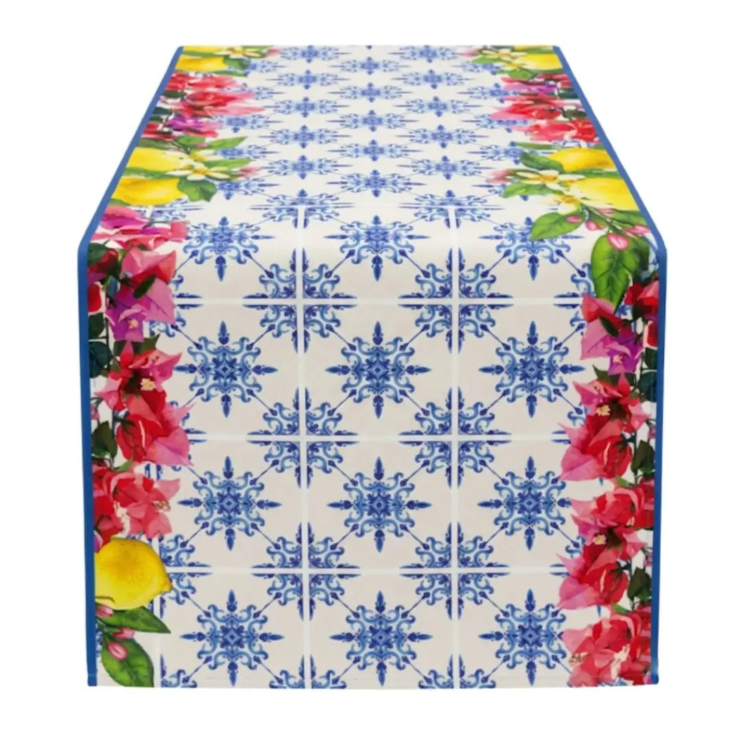 Runner con maioliche blu e fiori caratterizzato da un elegante motivo a piastrelle geometriche in stile mediterraneo su fondo bianco. I bordi di questo runner da tavola sono impreziositi da vivaci fiori rosa fucsia, limoni gialli e foglie verdi, ideali per decorare la tavola con freschezza.