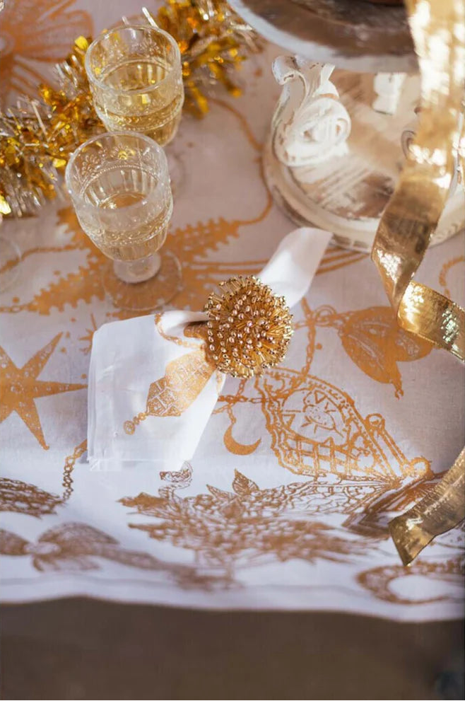Mise en place festiva con runner bianco e oro. Un tovagliolo bianco è impreziosito da un portatovagliolo a stella dorato. Sullo sfondo, calici di cristallo lavorato e decorazioni natalizie luminose.