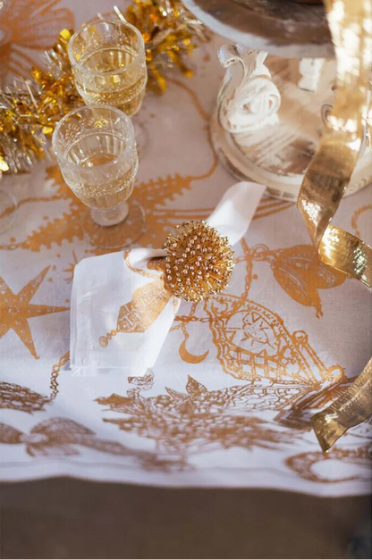 Mise en place festiva con runner bianco e oro. Un tovagliolo bianco è impreziosito da un portatovagliolo a stella dorato. Sullo sfondo, calici di cristallo lavorato e decorazioni natalizie luminose.