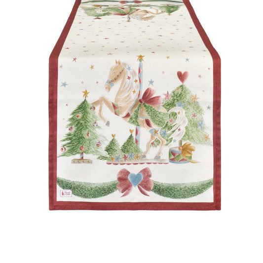 Runner natalizio in cotone Nuvole di Stoffa collezione "La Giostra di Amelie". Presenta un'illustrazione centrale con un cavallo bianco da giostra tra alberi di Natale addobbati, regali e stelline colorate su fondo panna. Il bordo è rifinito da una cornice rossa e una ghirlanda festiva con un cuore azzurro alla base.