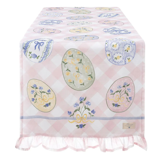 Runner da tavola della collezione "Pretty Easter" caratterizzato da un motivo a scacchi bianchi e rosa pastello con un'elegante gala arricciata sul bordo inferiore. Il tessuto è decorato con stampe di uova di Pasqua illustrate con roselline azzurre e fiocchi gialli, ideale per apparecchiare una tavola festiva romantica in perfetto stile shabby chic.