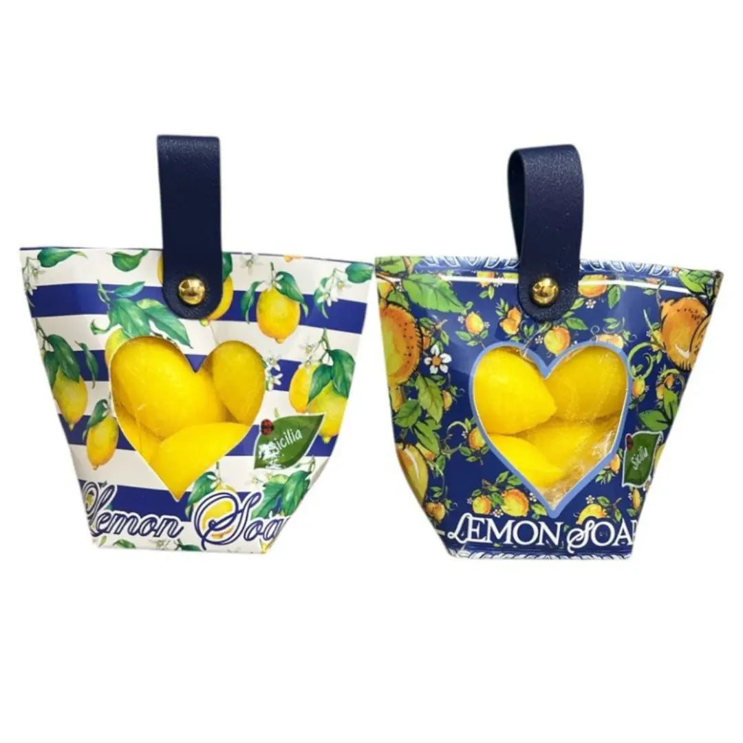 Saponetta al limone a forma di agrume custodita all'interno di una graziosa borsetta regalo assortita con manico in finta pelle blu firmata La Divina Costiera. Una confezione decorativa con finestra trasparente a forma di cuore e motivi ispirati ad Amalfi e alla Sicilia, ideale come profumata bomboniera o originale idea regalo per la cura del corpo.