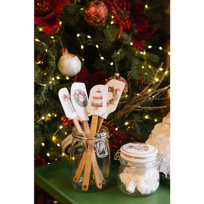 Spatole in silicone della collezione natalizia Nuvole di Stoffa esposte all'interno di un barattolo di vetro su un vassoio verde. Le spatole, con manico in legno, mostrano decorazioni di ghirlande, torte e pacchi regalo, immerse in un'atmosfera festiva con un albero di Natale addobbato e luci soffuse sullo sfondo.