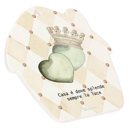 un elegante motivo a losanghe color crema e beige con piccoli pois marroni, al centro del quale spiccano i due cuori verde salvia e la corona regale. L'inquadratura di tre quarti permette di apprezzare il bordo bianco e pulito della porcellana, sottolineando la solidità dell'oggetto. La scritta "Casa è dove splende sempre la luce" è chiaramente leggibile nella parte inferiore, così come il foro funzionale sulla punta del tetto.