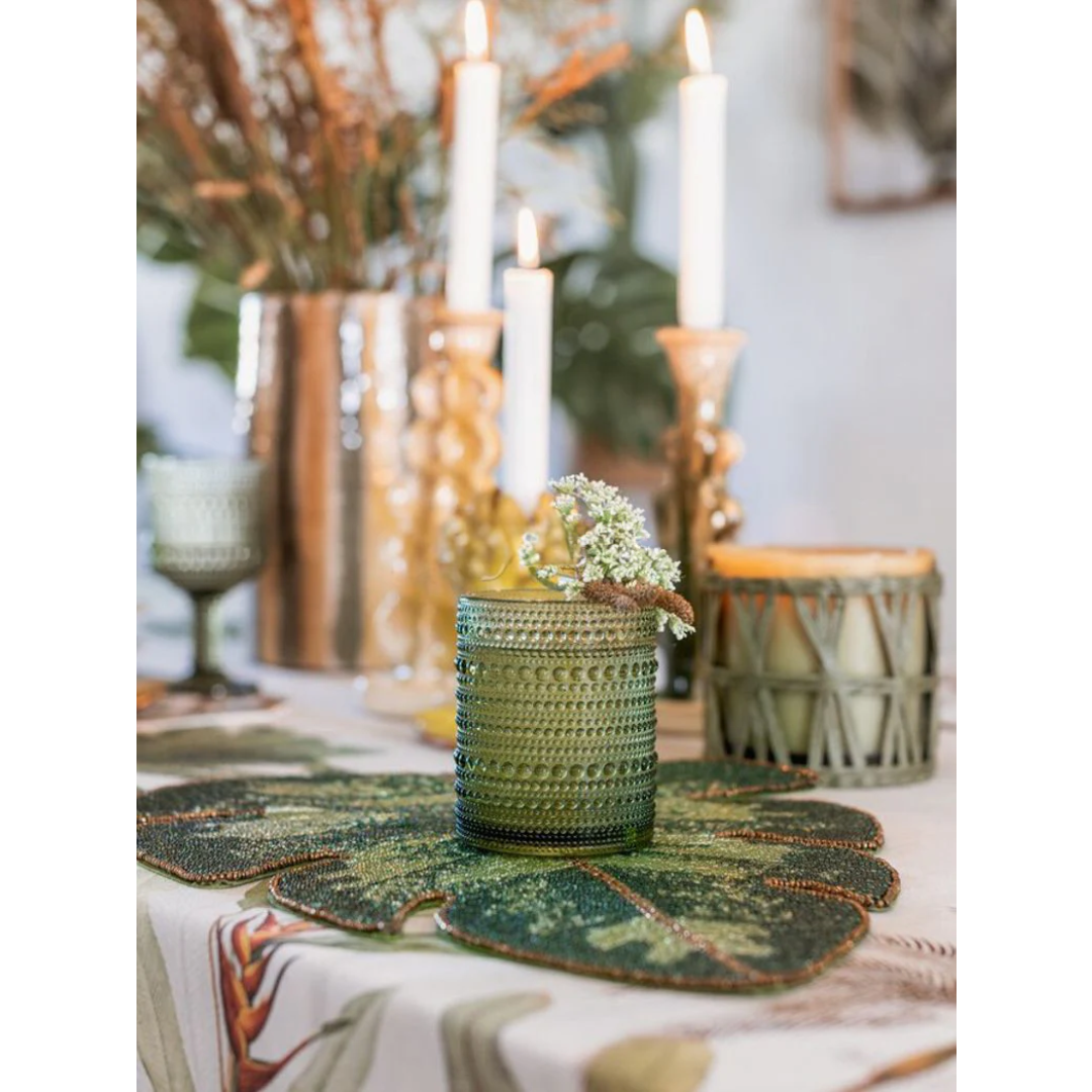 Mise en place elegante con sottopiatto a forma di foglia di Monstera in perline verdi, utilizzato come base per un bicchiere in vetro lavorato. Sullo sfondo, candele accese e decorazioni floreali creano un'atmosfera calda e raffinata.