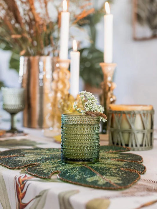 Mise en place elegante con sottopiatto a forma di foglia di Monstera in perline verdi, utilizzato come base per un bicchiere in vetro lavorato. Sullo sfondo, candele accese e decorazioni floreali creano un'atmosfera calda e raffinata.
