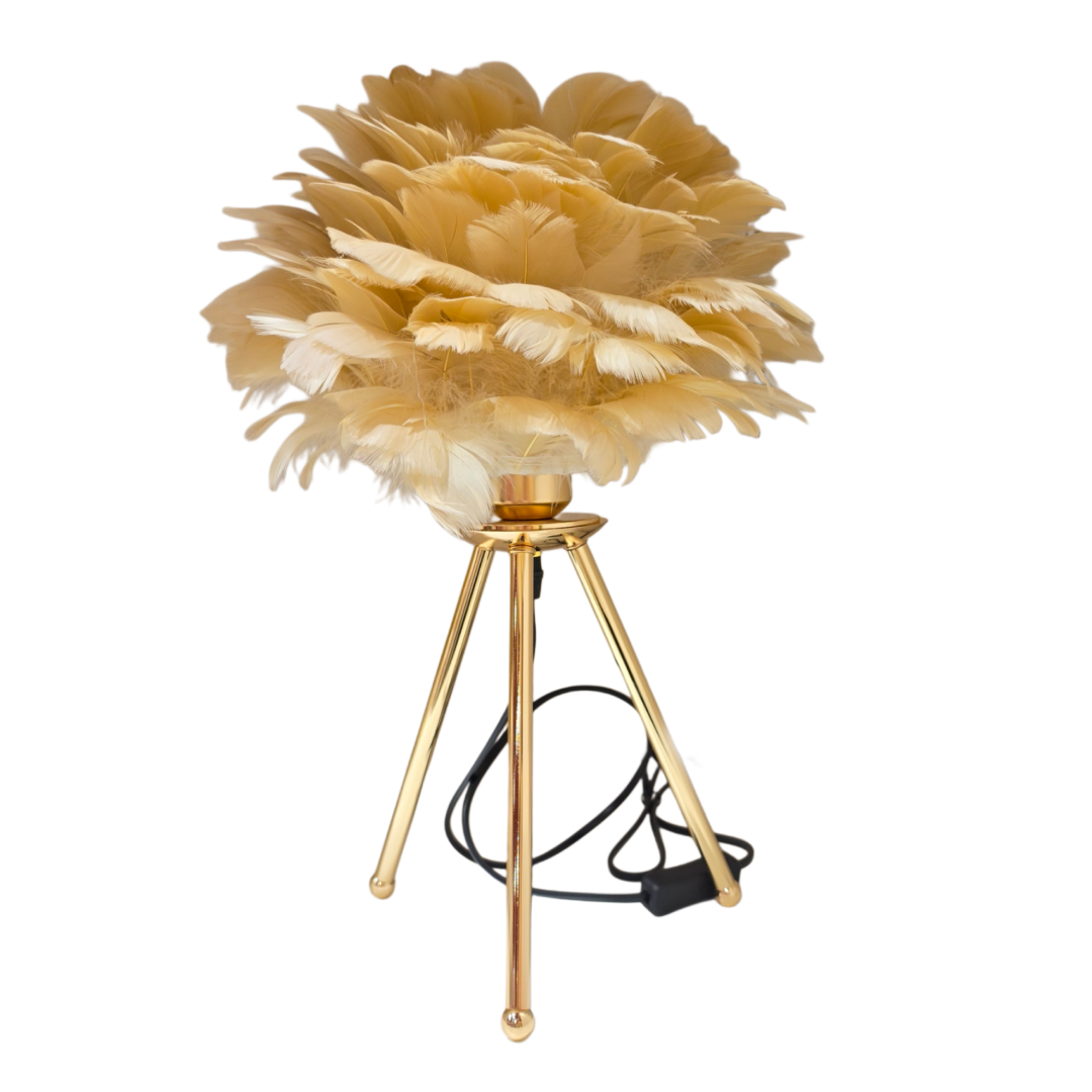 Lampada da tavolo con paralume realizzato in soffici piume beige e crema disposte a formare una nuvola voluminosa, sorretta da un treppiede in metallo dorato con cavo nero a vista. Lampada decorativa dal design glamour firmata Sud Import, ideale come complemento d'arredo per il comodino o come idea regalo raffinata.