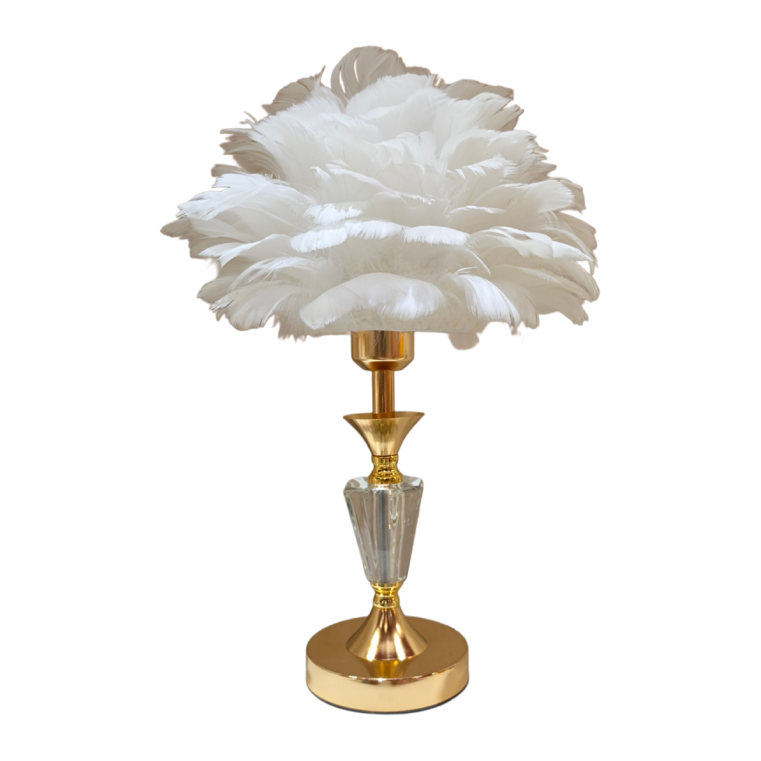 Lampada da tavolo con paralume realizzato in soffici piume panna e crema disposte a formare una nuvola voluminosa, sorretta da un treppiede in metallo dorato e vetro trasparente con cavo nero a vista. Lampada decorativa dal design glamour firmata Sud Import, ideale come complemento d'arredo per il comodino o come idea regalo raffinata.
