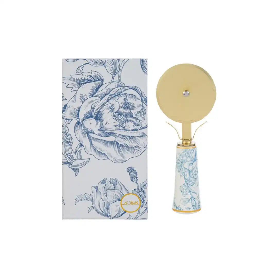 Tagliapizza con decoro azzurro firmato Le Stelle, caratterizzato da una lama rotante circolare in metallo color oro e un elegante manico in porcellana bianca decorato con delicati motivi floreali celestini. Questo raffinato utensile da cucina di design è accompagnato dalla sua custodia regalo rettangolare coordinata con la stessa fantasia botanica, perfetto come bomboniera di classe o idea regalo home decor.