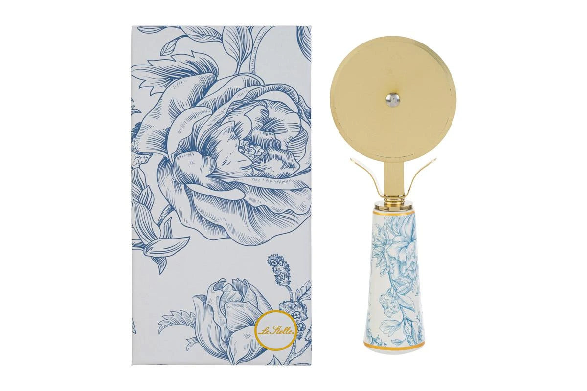 Tagliapizza con decoro azzurro firmato Le Stelle, caratterizzato da una lama rotante circolare in metallo color oro e un elegante manico in porcellana bianca decorato con delicati motivi floreali celestini. Questo raffinato utensile da cucina di design è accompagnato dalla sua custodia regalo rettangolare coordinata con la stessa fantasia botanica, perfetto come bomboniera di classe o idea regalo home decor.