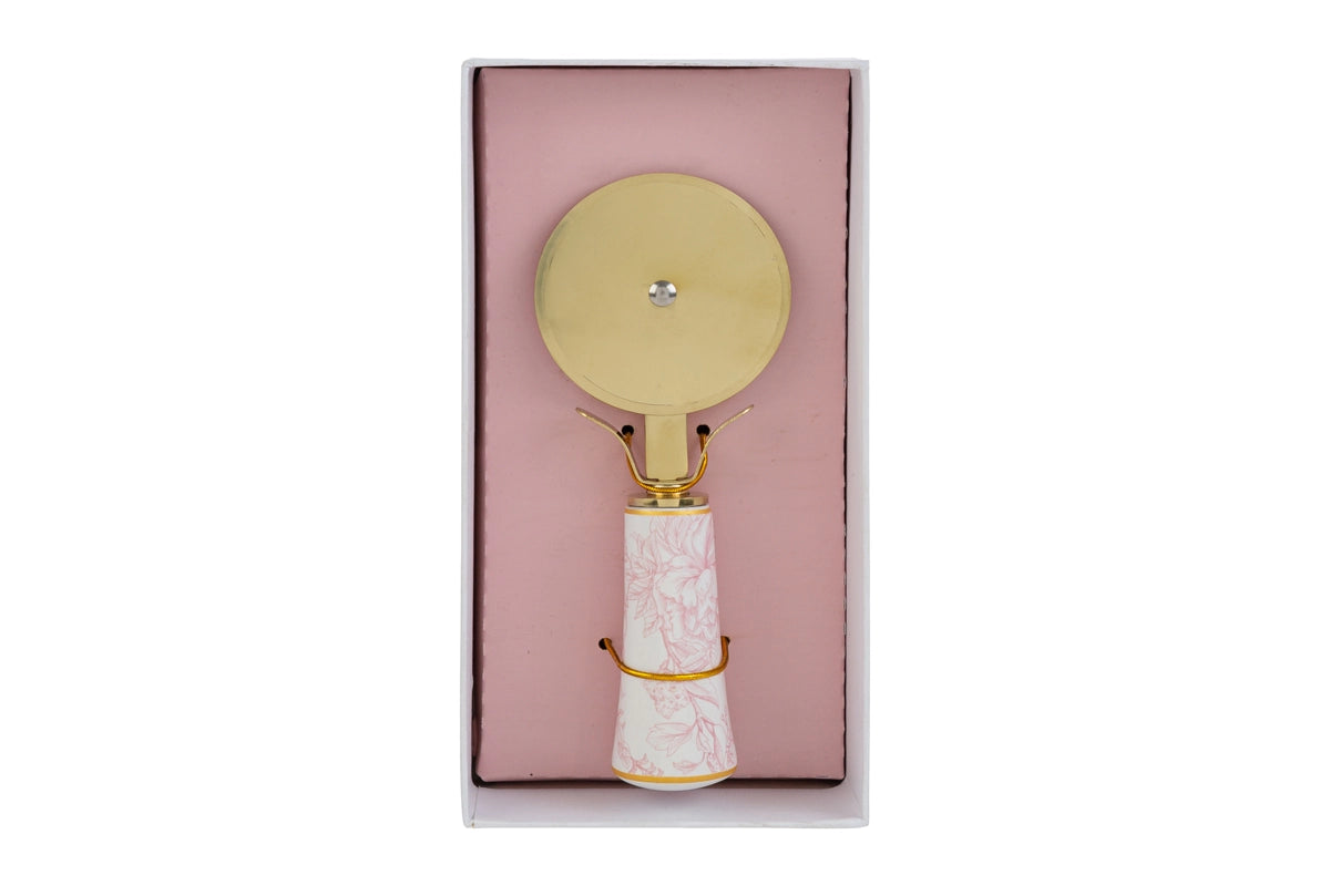 Tagliapizza con decoro rosa firmato Le Stelle, caratterizzato da una lama rotante circolare in metallo color oro e un elegante manico in porcellana bianca decorato con delicati motivi floreali rosati. Questo raffinato utensile da cucina di design è accompagnato dalla sua custodia regalo rettangolare coordinata con la stessa fantasia botanica, perfetto come bomboniera di classe o idea regalo home decor.