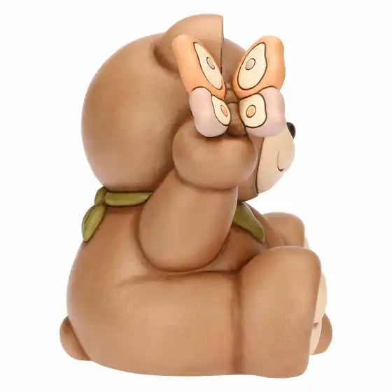 Statuina maxi in ceramica dipinta a mano raffigurante il classico orsetto Teddy di Thun. L'orso è seduto in posa sorridente con un occhio chiuso in un occhiolino, indossa un fazzoletto verde al collo e tiene una farfalla rosa e arancione appoggiata sull'orecchio destro sollevato. Design caratterizzato dalle tipiche forme morbide e dai colori caldi del brand.