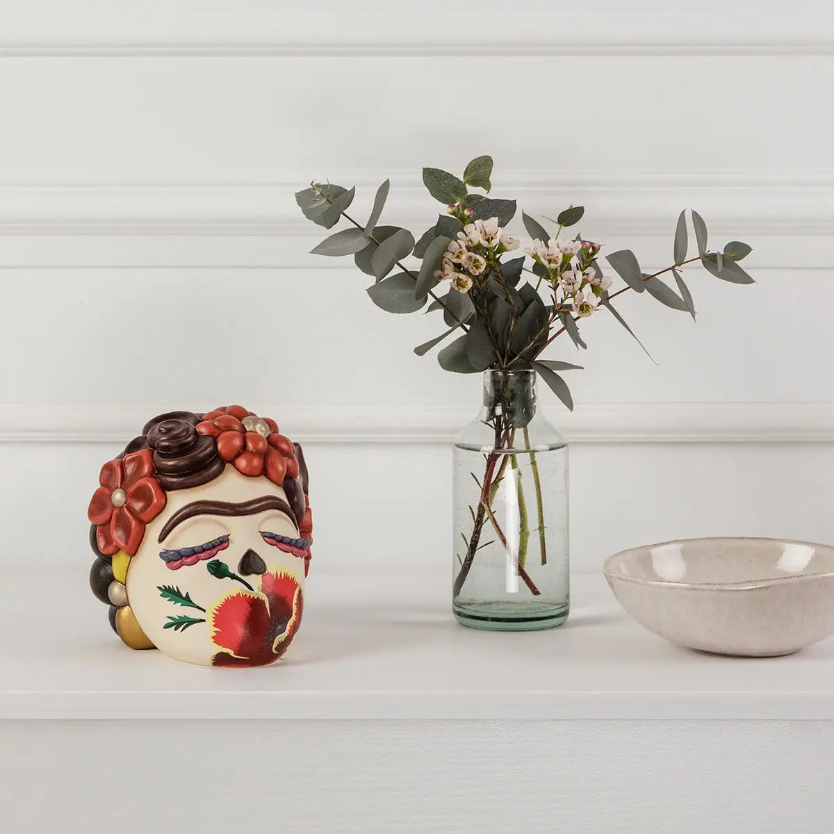 Elegante ambientazione d'interni che mostra la testa decorativa Frida di Thun esposta su un ripiano bianco minimalista. La scultura in ceramica, con i suoi vivaci fiori rossi e il decoro a papavero, è affiancata da un raffinato vaso in vetro trasparente con rami di eucalipto e piccoli fiori bianchi, creando un angolo d'arredo armonioso e di tendenza.
