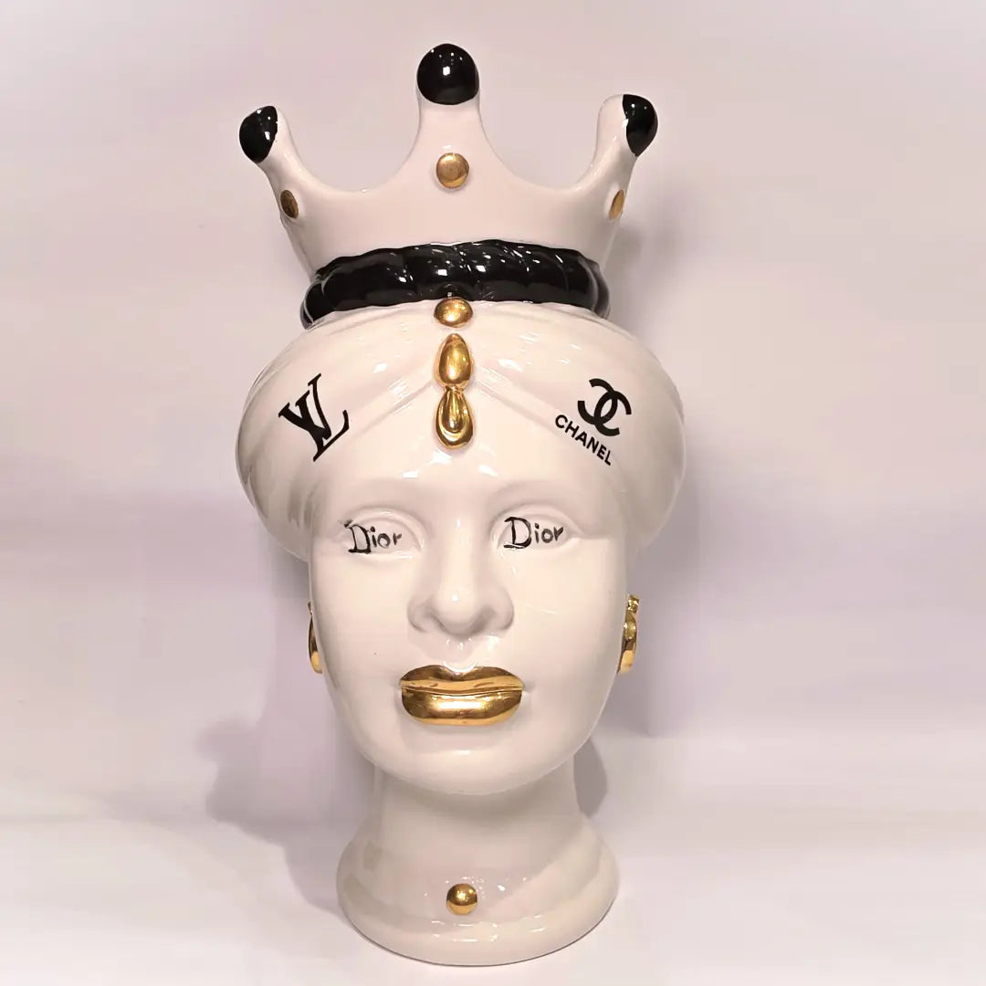 Coppia di Teste di Moro in ceramica bianca lucida con turbanti neri e bianchi decorati da loghi fashion neri e dorati, completate da corone regali e labbra con finitura in oro zecchino. Queste sculture ornamentali di design pop art fondono la tradizione ceramica siciliana con l'estetica del lusso moderno, rappresentando un complemento d'arredo audace ed esclusivo per interni ricercati.