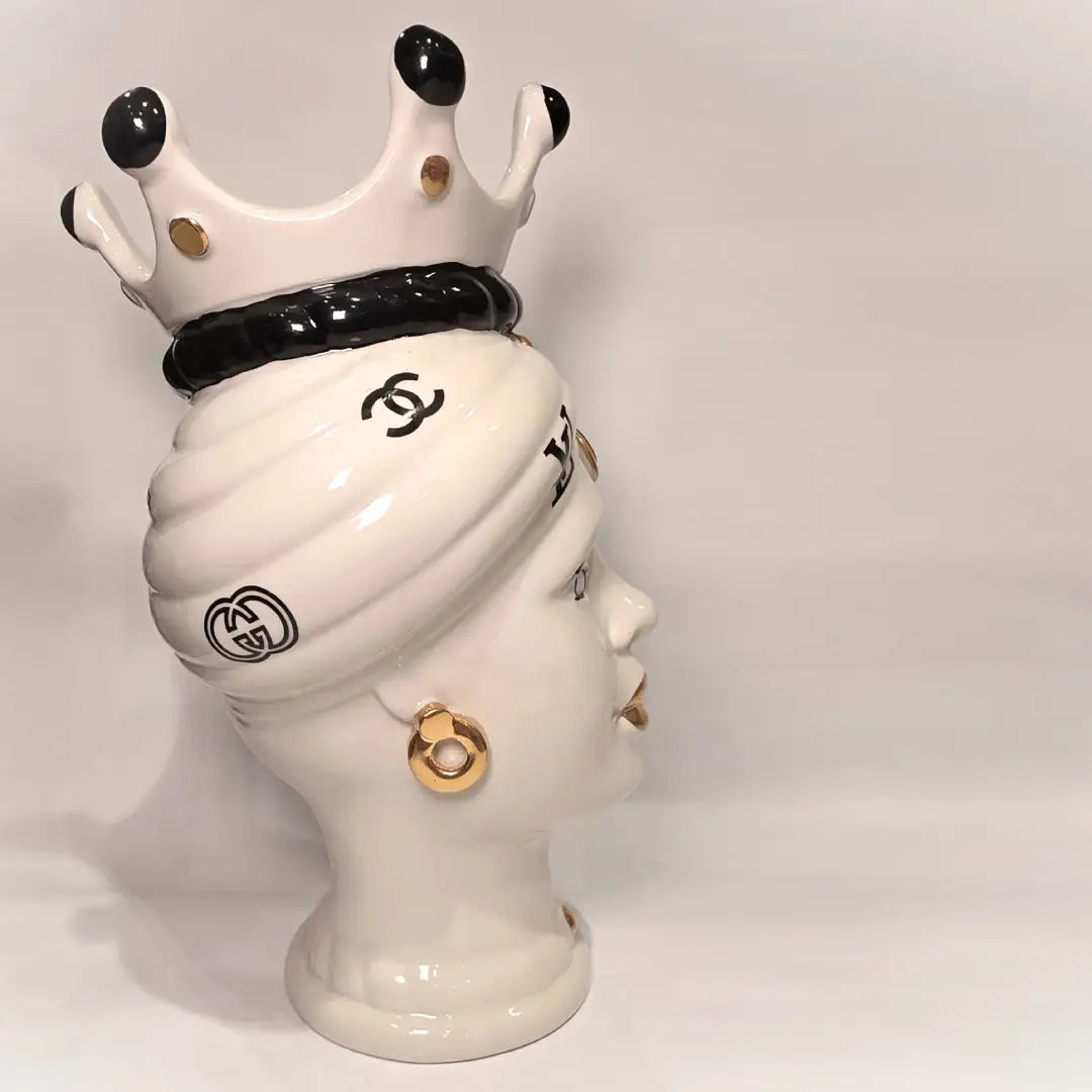 Coppia di Teste di Moro in ceramica bianca lucida con turbanti neri e bianchi decorati da loghi fashion neri e dorati, completate da corone regali e labbra con finitura in oro zecchino. Queste sculture ornamentali di design pop art fondono la tradizione ceramica siciliana con l'estetica del lusso moderno, rappresentando un complemento d'arredo audace ed esclusivo per interni ricercati.