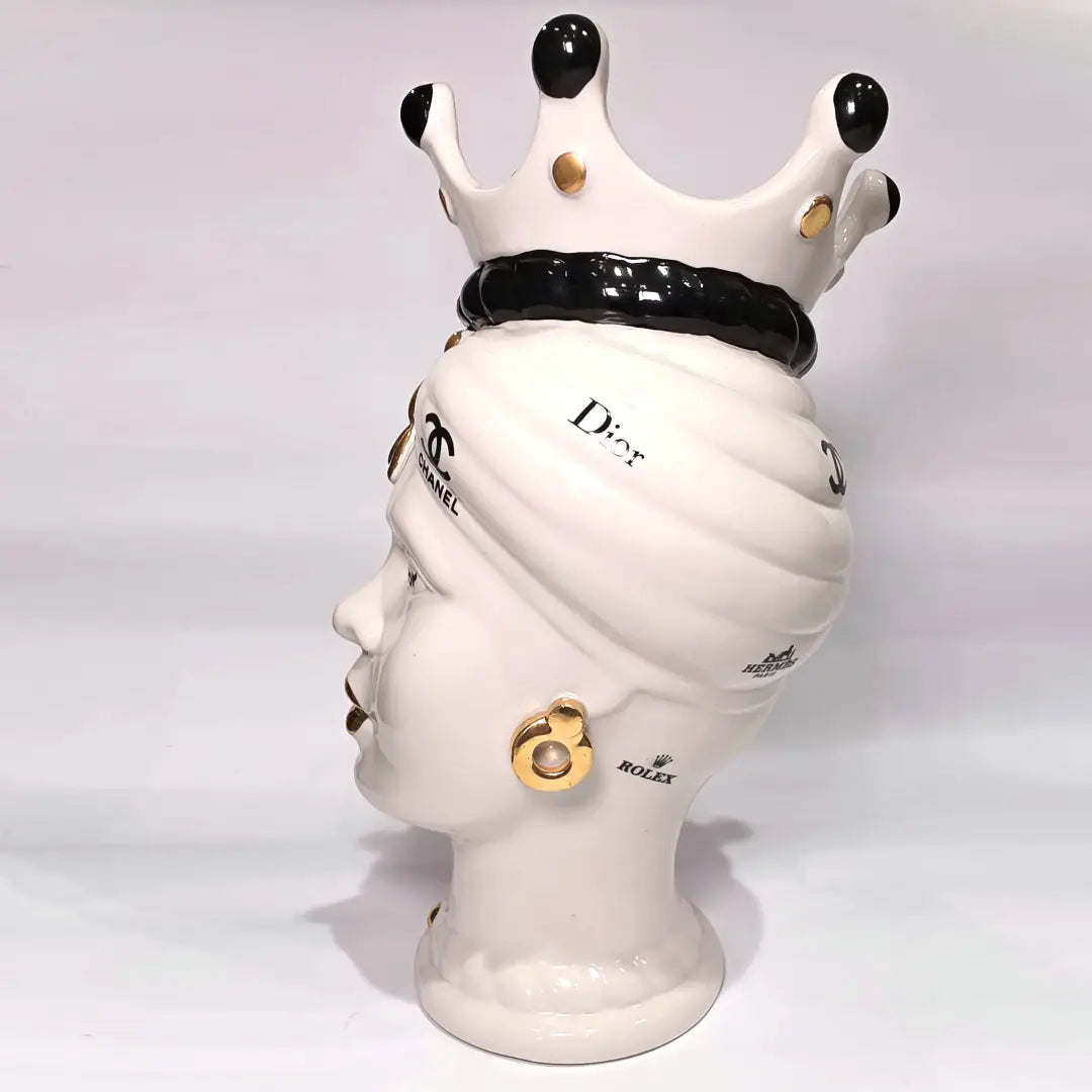 Coppia di Teste di Moro in ceramica bianca lucida con turbanti neri e bianchi decorati da loghi fashion neri e dorati, completate da corone regali e labbra con finitura in oro zecchino. Queste sculture ornamentali di design pop art fondono la tradizione ceramica siciliana con l'estetica del lusso moderno, rappresentando un complemento d'arredo audace ed esclusivo per interni ricercati.