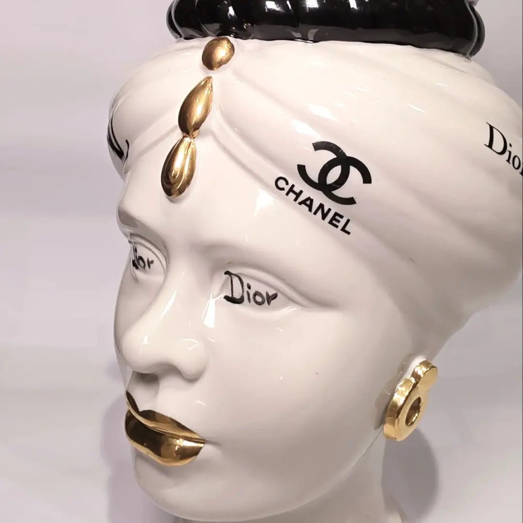 Coppia di Teste di Moro in ceramica bianca lucida con turbanti neri e bianchi decorati da loghi fashion neri e dorati, completate da corone regali e labbra con finitura in oro zecchino. Queste sculture ornamentali di design pop art fondono la tradizione ceramica siciliana con l'estetica del lusso moderno, rappresentando un complemento d'arredo audace ed esclusivo per interni ricercati.