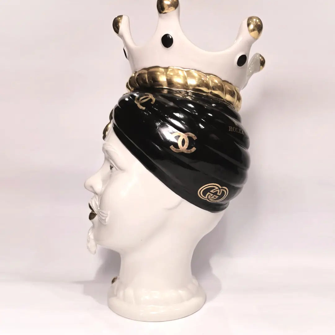 Coppia di Teste di Moro in ceramica bianca lucida con turbanti neri e bianchi decorati da loghi fashion neri e dorati, completate da corone regali e labbra con finitura in oro zecchino. Queste sculture ornamentali di design pop art fondono la tradizione ceramica siciliana con l'estetica del lusso moderno, rappresentando un complemento d'arredo audace ed esclusivo per interni ricercati.