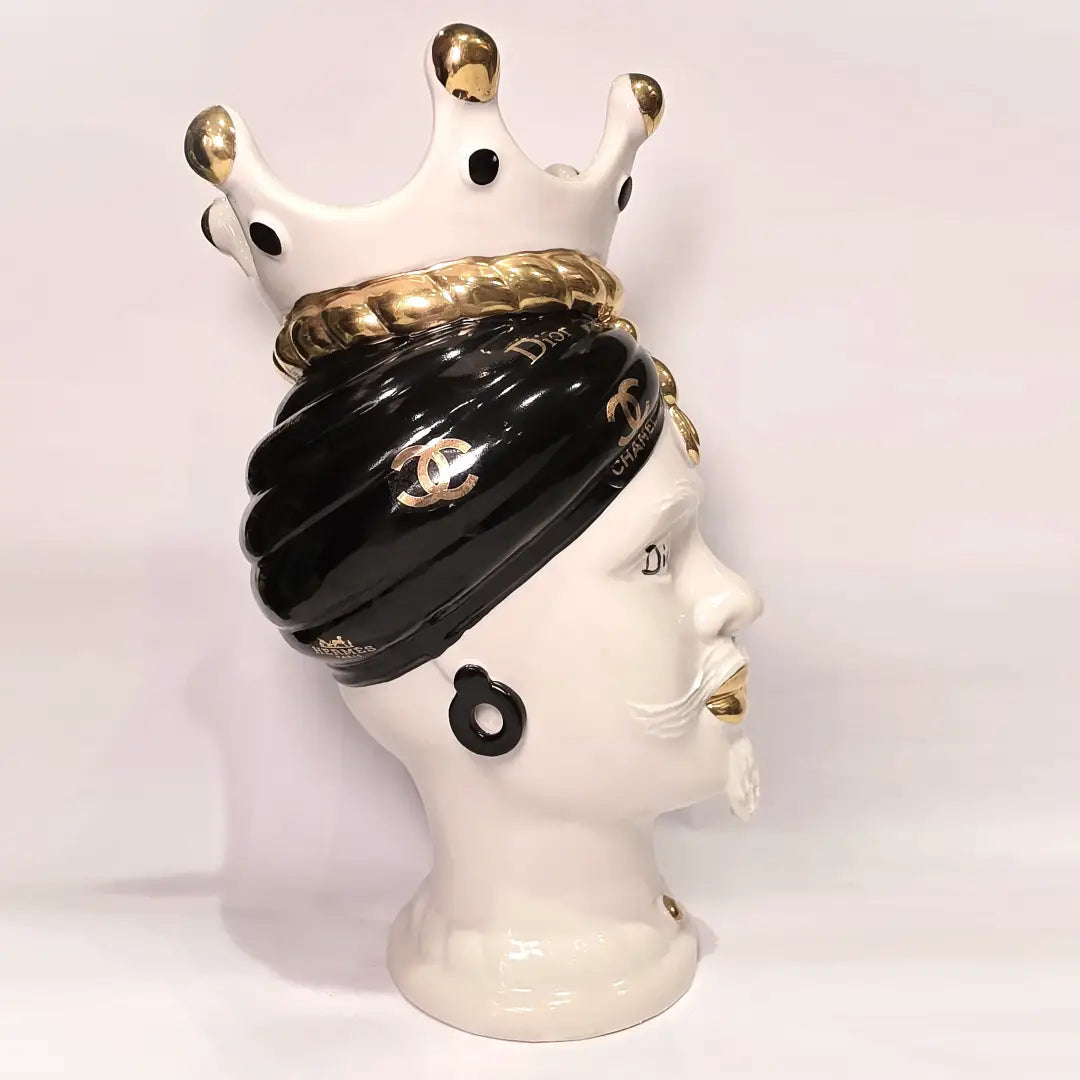 Coppia di Teste di Moro in ceramica bianca lucida con turbanti neri e bianchi decorati da loghi fashion neri e dorati, completate da corone regali e labbra con finitura in oro zecchino. Queste sculture ornamentali di design pop art fondono la tradizione ceramica siciliana con l'estetica del lusso moderno, rappresentando un complemento d'arredo audace ed esclusivo per interni ricercati.
