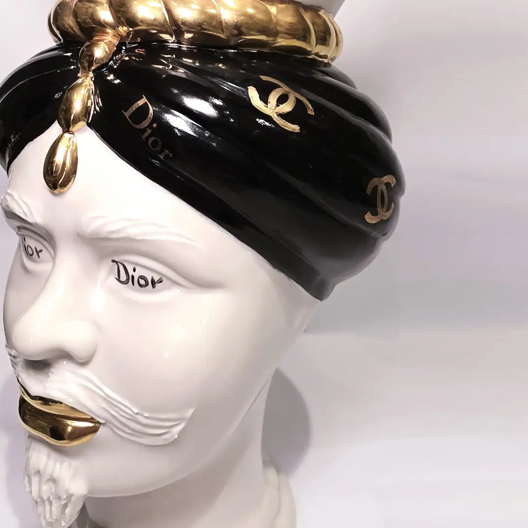 Coppia di Teste di Moro in ceramica bianca lucida con turbanti neri e bianchi decorati da loghi fashion neri e dorati, completate da corone regali e labbra con finitura in oro zecchino. Queste sculture ornamentali di design pop art fondono la tradizione ceramica siciliana con l'estetica del lusso moderno, rappresentando un complemento d'arredo audace ed esclusivo per interni ricercati.
