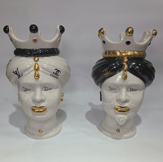 Coppia di Teste di Moro in ceramica bianca lucida con turbanti neri e bianchi decorati da loghi fashion neri e dorati, completate da corone regali e labbra con finitura in oro zecchino. Queste sculture ornamentali di design pop art fondono la tradizione ceramica siciliana con l'estetica del lusso moderno, rappresentando un complemento d'arredo audace ed esclusivo per interni ricercati.