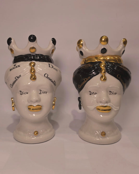 Coppia di Teste di Moro in ceramica bianca lucida con turbanti neri e bianchi decorati da loghi fashion neri e dorati, completate da corone regali e labbra con finitura in oro zecchino. Queste sculture ornamentali di design pop art fondono la tradizione ceramica siciliana con l'estetica del lusso moderno, rappresentando un complemento d'arredo audace ed esclusivo per interni ricercati.