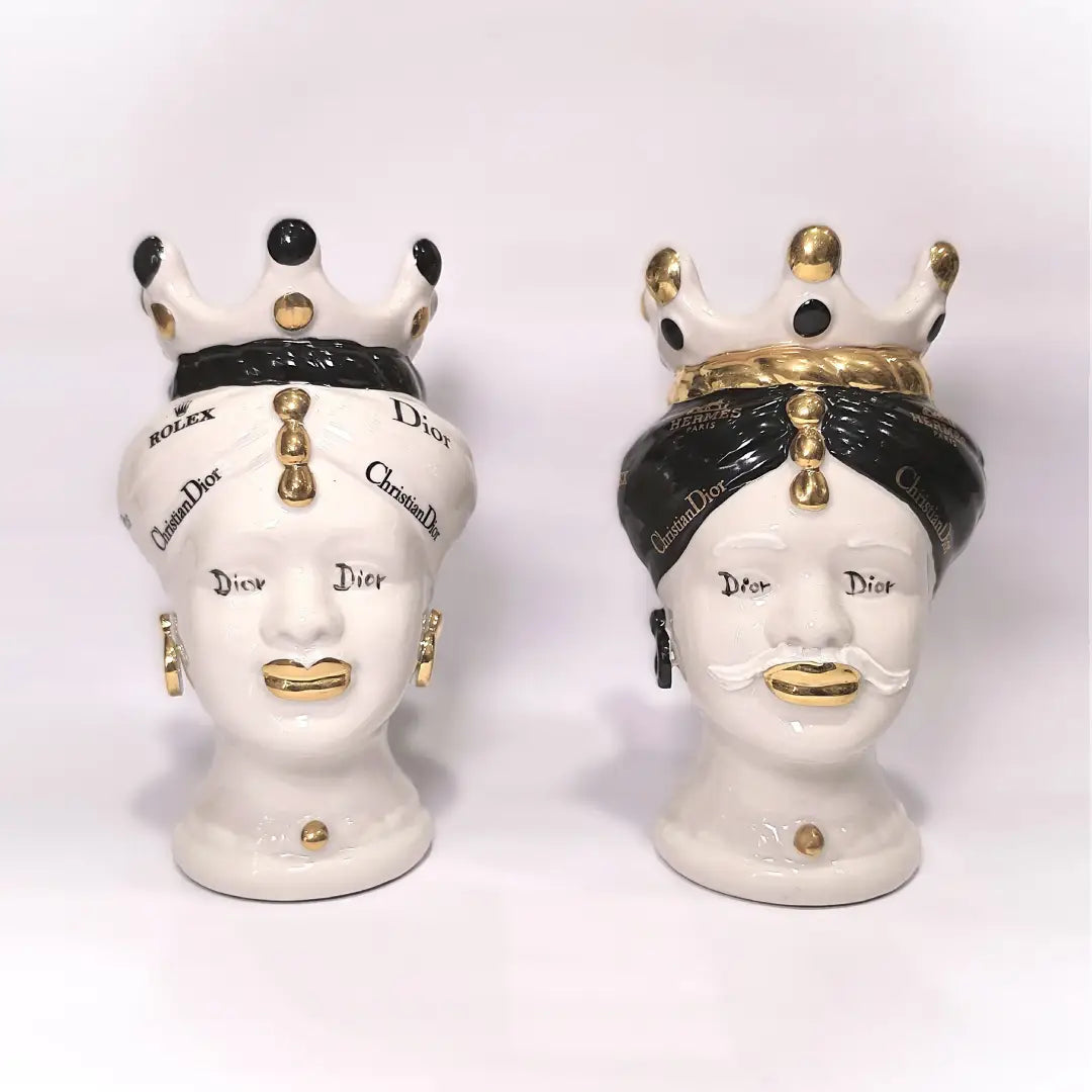 Coppia di Teste di Moro in ceramica bianca lucida con turbanti neri e bianchi decorati da loghi fashion neri e dorati, completate da corone regali e labbra con finitura in oro zecchino. Queste sculture ornamentali di design pop art fondono la tradizione ceramica siciliana con l'estetica del lusso moderno, rappresentando un complemento d'arredo audace ed esclusivo per interni ricercati.