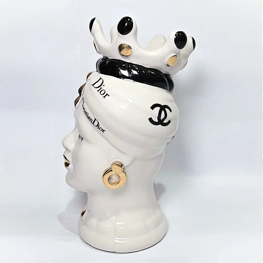 Coppia di Teste di Moro in ceramica bianca lucida con turbanti neri e bianchi decorati da loghi fashion neri e dorati, completate da corone regali e labbra con finitura in oro zecchino. Queste sculture ornamentali di design pop art fondono la tradizione ceramica siciliana con l'estetica del lusso moderno, rappresentando un complemento d'arredo audace ed esclusivo per interni ricercati.