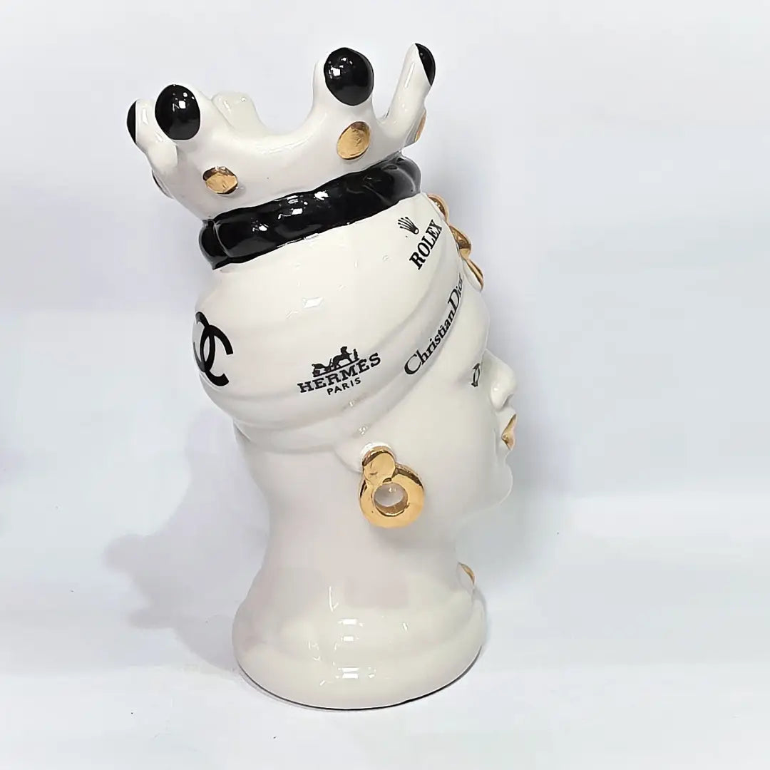 Coppia di Teste di Moro in ceramica bianca lucida con turbanti neri e bianchi decorati da loghi fashion neri e dorati, completate da corone regali e labbra con finitura in oro zecchino. Queste sculture ornamentali di design pop art fondono la tradizione ceramica siciliana con l'estetica del lusso moderno, rappresentando un complemento d'arredo audace ed esclusivo per interni ricercati.
