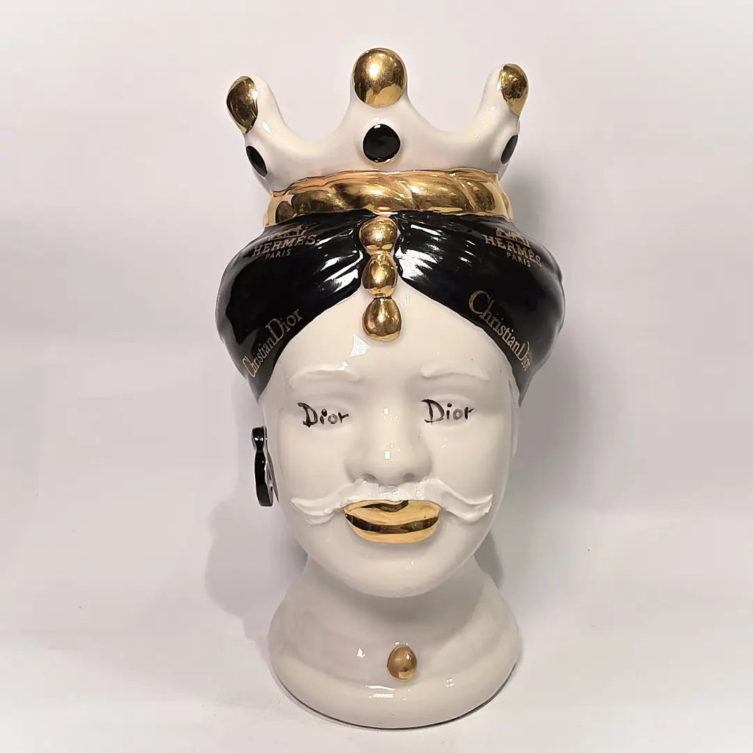Coppia di Teste di Moro in ceramica bianca lucida con turbanti neri e bianchi decorati da loghi fashion neri e dorati, completate da corone regali e labbra con finitura in oro zecchino. Queste sculture ornamentali di design pop art fondono la tradizione ceramica siciliana con l'estetica del lusso moderno, rappresentando un complemento d'arredo audace ed esclusivo per interni ricercati.