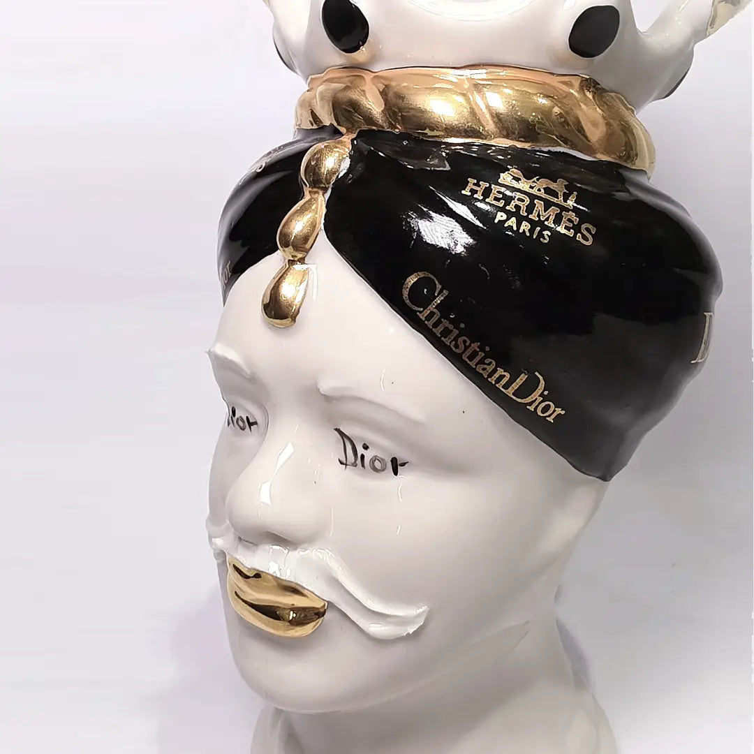 Coppia di Teste di Moro in ceramica bianca lucida con turbanti neri e bianchi decorati da loghi fashion neri e dorati, completate da corone regali e labbra con finitura in oro zecchino. Queste sculture ornamentali di design pop art fondono la tradizione ceramica siciliana con l'estetica del lusso moderno, rappresentando un complemento d'arredo audace ed esclusivo per interni ricercati.
