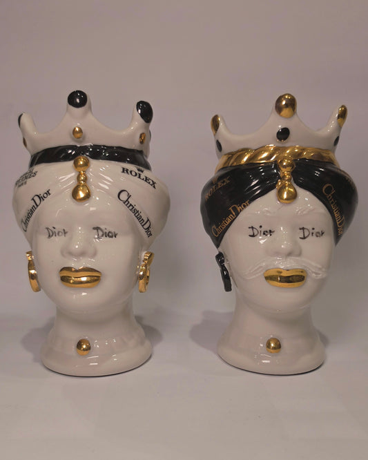 Coppia di Teste di Moro in ceramica bianca lucida con turbanti neri e bianchi decorati da loghi fashion neri e dorati, completate da corone regali e labbra con finitura in oro zecchino. Queste sculture ornamentali di design pop art fondono la tradizione ceramica siciliana con l'estetica del lusso moderno, rappresentando un complemento d'arredo audace ed esclusivo per interni ricercati.