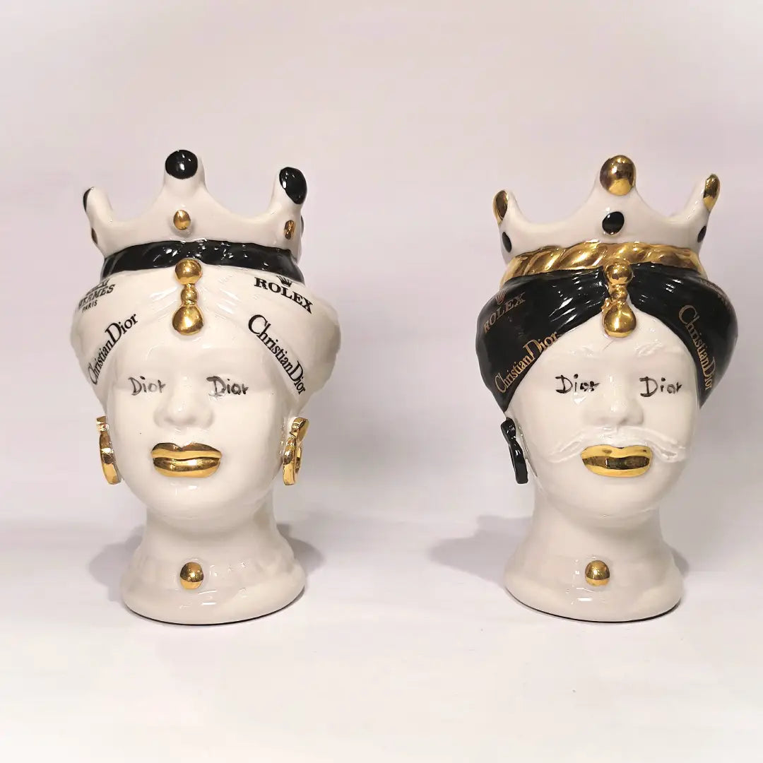 Coppia di Teste di Moro in ceramica bianca lucida con turbanti neri e bianchi decorati da loghi fashion neri e dorati, completate da corone regali e labbra con finitura in oro zecchino. Queste sculture ornamentali di design pop art fondono la tradizione ceramica siciliana con l'estetica del lusso moderno, rappresentando un complemento d'arredo audace ed esclusivo per interni ricercati.